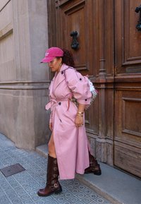 Trench coat rosa con bottoni neri, una cintura marrone e stivali al ginocchio marroni con fibbie. Gli accessori includono un cappello rosa e uno zaino a fantasia.