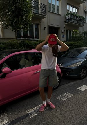 Joven con camiseta blanca, pantalones cortos grises y zapatillas rosas ajusta una gorra roja mientras está de pie junto a un coche rosa en una calle de la ciudad con edificios.