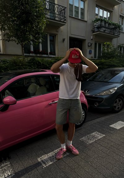 Joven con camiseta blanca, pantalones cortos grises y zapatillas rosas ajusta una gorra roja mientras está de pie junto a un coche rosa en una calle de la ciudad con edificios.