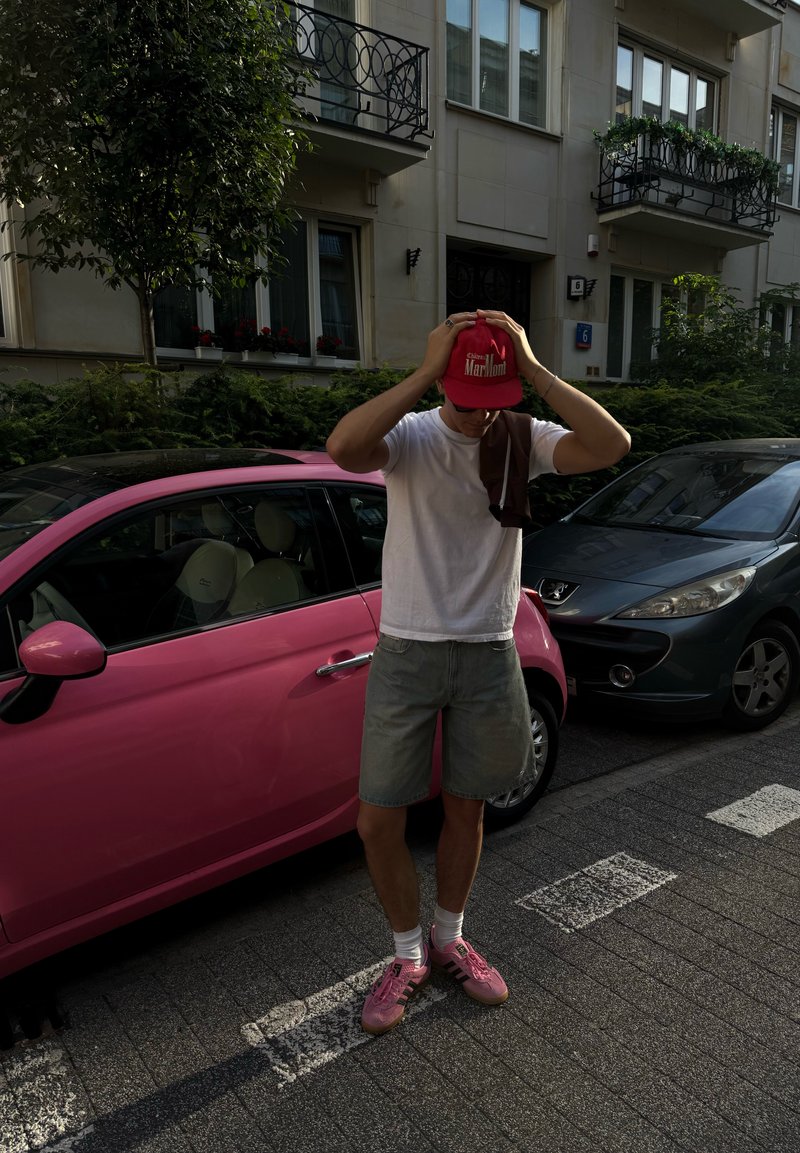 Giovane uomo con maglietta bianca, pantaloni corti grigi e scarpe da ginnastica rosa sistemando un cappellino rosso mentre si trova accanto a un'auto rosa su una strada di città con edifici.