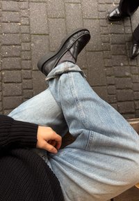 Črne usnjene čevlje s okroglo konico, v kombinaciji z svetloblue jeansom in črnim pletenim puloverjem. Okolje vključuje tlakovane ulice.