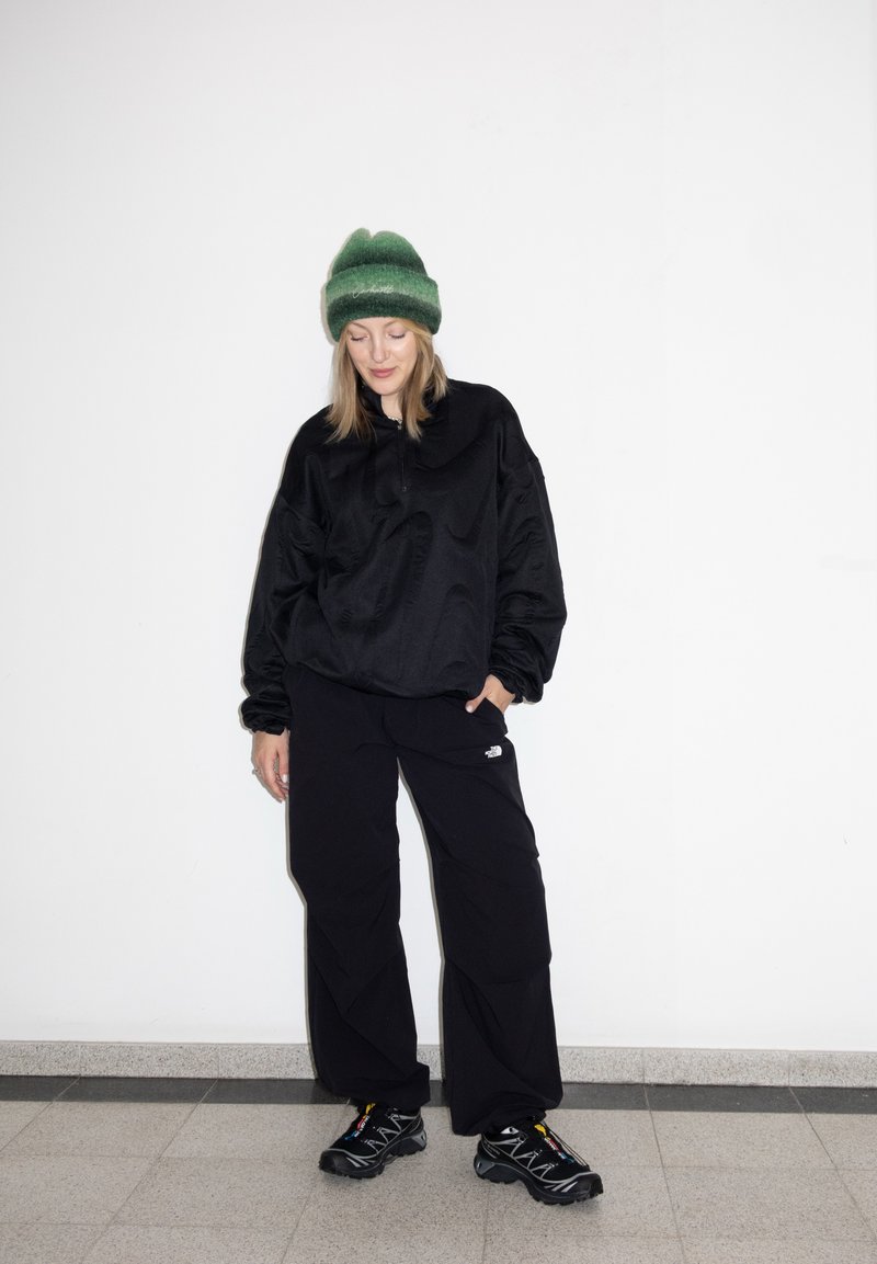 Femme portant un bonnet vert, une veste noire oversize, un pantalon noir North Face et des baskets noires, debout contre un mur blanc.