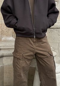 Sweat à capuche gris foncé zippé avec poches avant, associé à un pantalon cargo marron clair avec plusieurs poches latérales et un tissu texturé.