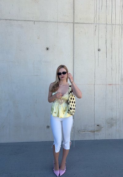 Mujer con gafas de sol, top de satén amarillo, pantalones blancos cortos, tacones rosas y un bolso de hombro con lunares, de pie junto a una pared de hormigón.