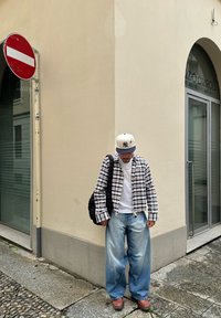 Camicia a scacchi bianca e nera, t-shirt bianca, jeans blu larghi, scarpe marroni e un cappellino beige, in piedi vicino a un muro all'angolo.