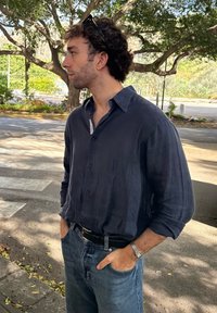 Granatowa koszula z krótkim rękawem, lekka w dotyku, noszona z niebieskimi dżinsami i czarnym paskiem. Ręce w kieszeniach, srebrny zegarek na nadgarstku.