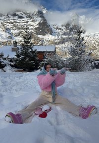 Personne portant une veste rose et une écharpe bleue assise dans la neige, jambes écartées, jetant de la neige en l'air, avec une montagne enneigée et des arbres en arrière-plan.