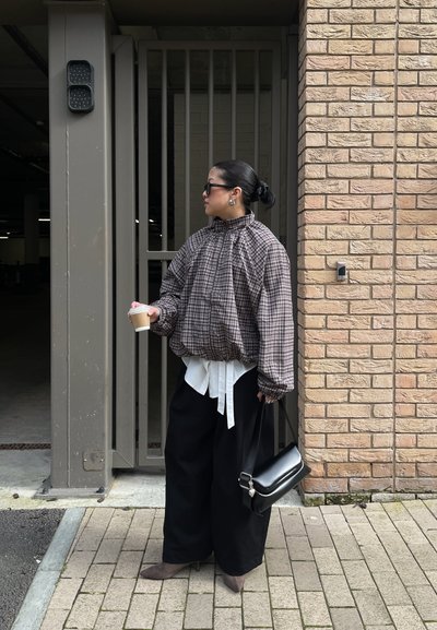 Mujer con moño, que lleva gafas de sol, chaqueta de cuadros, pantalones negros anchos, sosteniendo una taza de café y un bolso negro, de pie contra una pared de ladrillos.