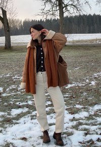Femme debout à l'extérieur en tenue d'hiver, portant un pantalon beige, des bottes noires, une chemise noire, une veste en peau retournée marron et une écharpe couleur rouille, touchant son oreille.