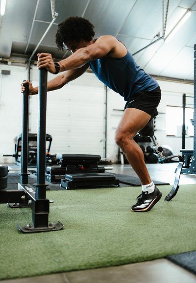 Atleta utilizando un empuje de trineo en un gimnasio, vistiendo una camiseta sin mangas azul y pantalones cortos negros, con una pierna prostética de fibra de carbono y zapatos de entrenamiento negros.