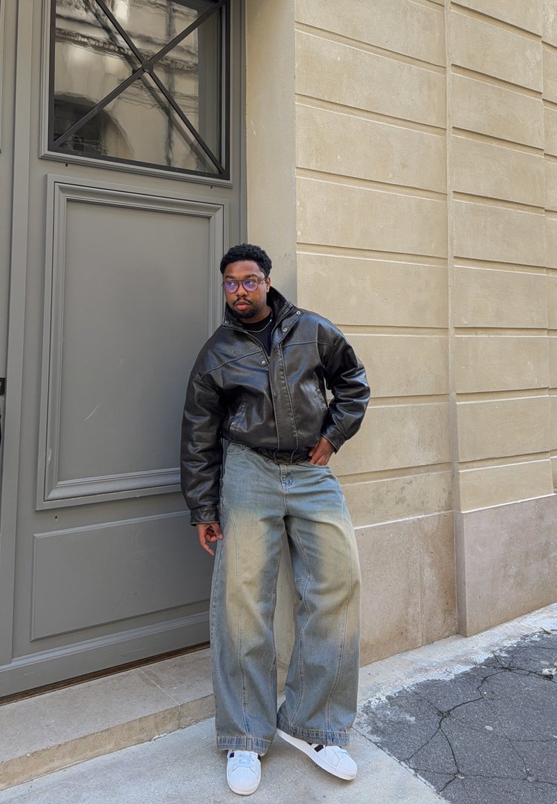 Homme portant une veste en cuir noire, un jean large bleu clair, des baskets blanches et des lunettes, appuyé contre un mur de pierre beige près d'une porte grise.