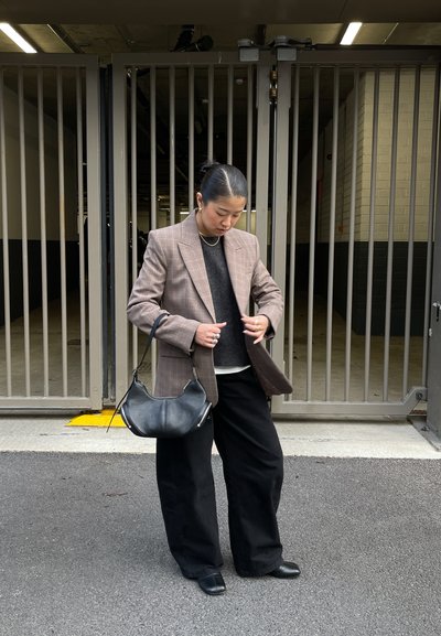 Blazer de cuadros marrón sobre un suéter oscuro, combinado con pantalones negros de pierna ancha y un bolso negro de hombro. Tela lisa con diseño entallado.