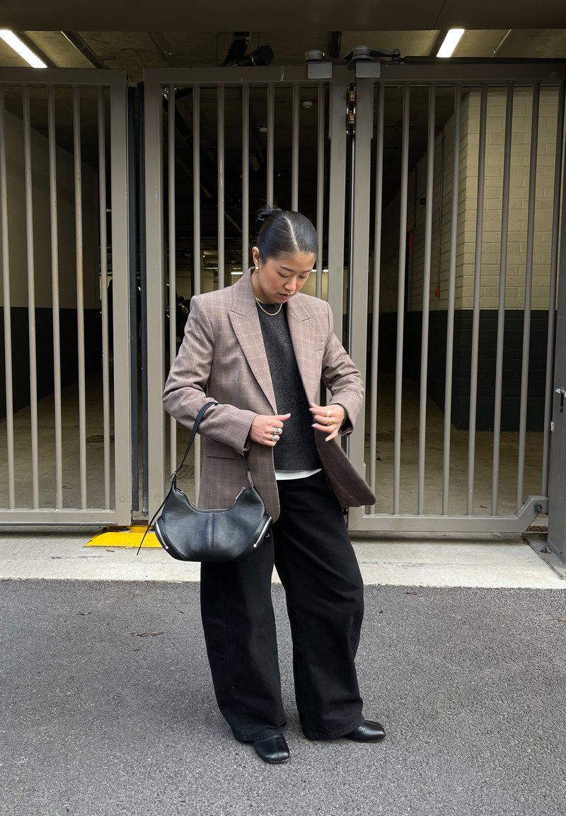 Blazer à carreaux marron sur un pull sombre, associé à un pantalon large noir et un sac à main noir. Tissu lisse avec une coupe ajustée.
