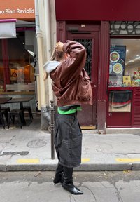 Person som justerer håret, iført brun lærjakke, grått skjørt og svarte spisse støvler, står på byfortauet ved siden av en rød butikkfasade.