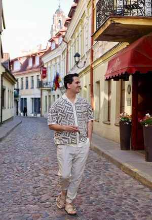 Camisa de manga corta con un patrón geométrico en beige y marrón, pantalones beiges y sandalias. Calle de adoquines con edificios coloridos.