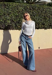 Haut blanc à manches longues avec un col bénitier, jean en denim bleu à jambes larges, et un sac à main bordeaux. Texture lisse, coupe décontractée, et couleurs neutres.