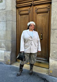 Blazer supradimensionat bej deschis, top alb, pantaloni scurți verde camuflaj, cizme maro ciocolatiu până la genunchi, geantă neagră, cu baretă albă și ochelari de soare.