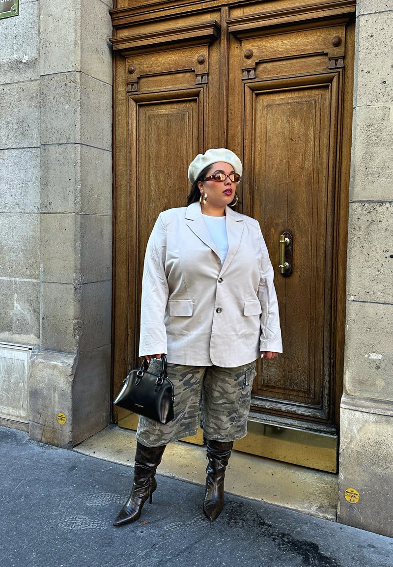 Veste surdimensionnée beige clair, haut blanc, shorts camouflage verts, bottes montantes chocolat, sac à main noir, avec un béret blanc et des lunettes de soleil.