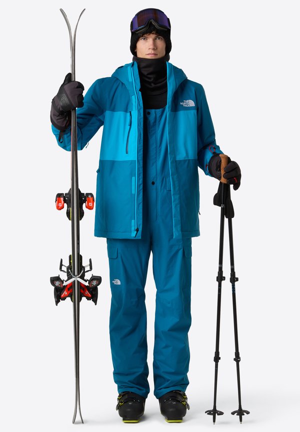 Blue ski jacket paired with matching trousers, gloves, and goggles. Holds skis featuring red bindings and black poles with cork grips. Black ski boots.