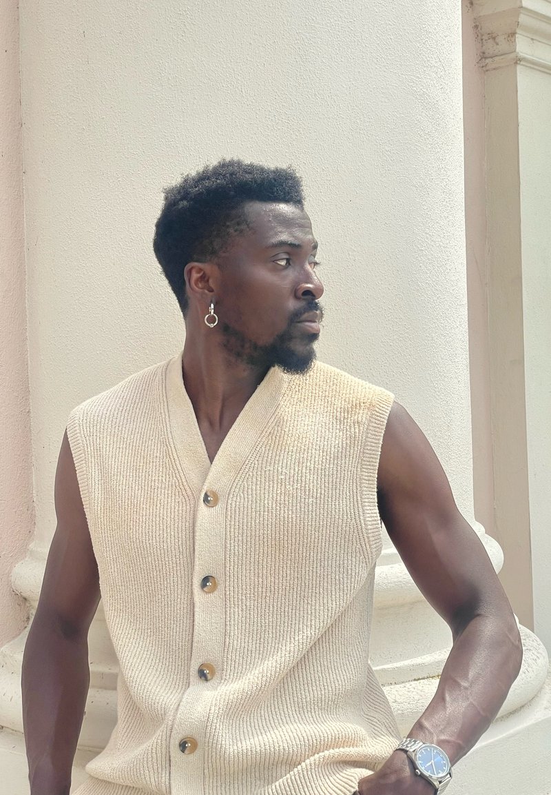 Gilet sans manches en tricot côtelé beige avec six boutons, porté sur un t-shirt. Présente un motif texturé et accessoirisé avec une boucle d'oreille en argent.