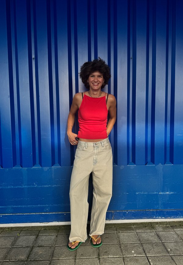 Donna sorridente con capelli corti e ricci, indossa una canottiera rossa, pantaloni beige a gamba larga e infradito verdi, in piedi contro un muro di metallo ondulato blu.