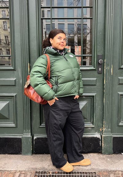 Mujer con chaqueta acolchada verde, pantalones negros y zapatos marrones, de pie con las manos en los bolsillos frente a grandes puertas dobles verdes.