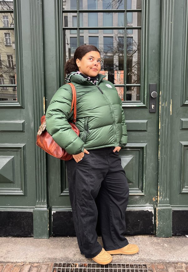 Kvinde iført en grøn dunjakke, sorte bukser og brune sko står med hænderne i lommerne foran store grønne dobbeltdøre.