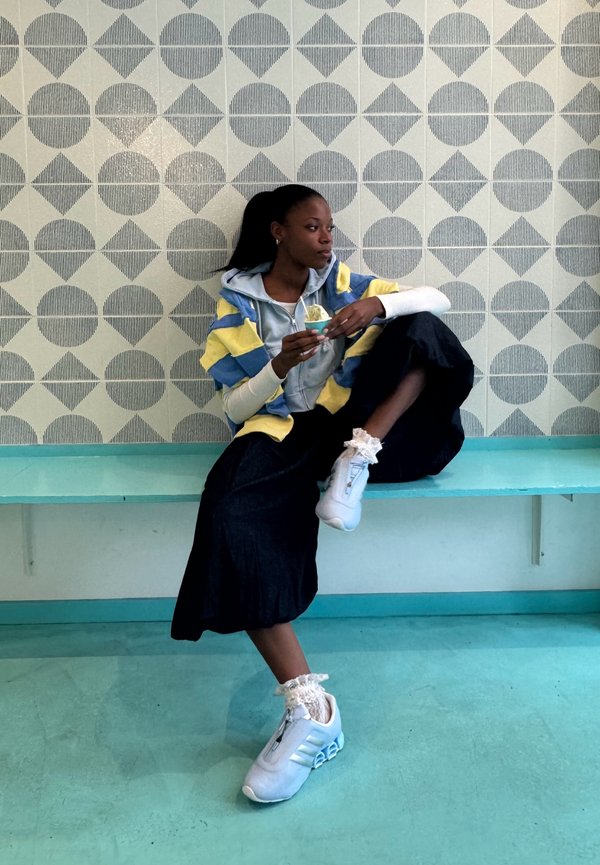 Young woman sitting on turquoise bench, eating ice cream, wearing black skirt, striped jacket, white sneakers, with geometric patterned wall behind.