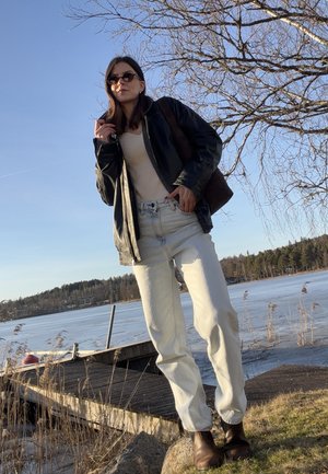 Mujer con gafas de sol, chaqueta de cuero negra, top beige y pantalones blancos, de pie junto a un muelle junto al lago con ramas de árboles desnudas y agua congelada.