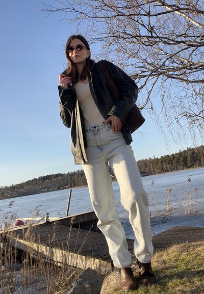 Mujer con gafas de sol, chaqueta de cuero negra, top beige y pantalones blancos, de pie junto a un muelle junto al lago con ramas de árboles desnudas y agua congelada.