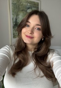 Portrett av en person med langt, bølget brunt hår som har på seg en hvit ribbestrikket genser. De har blå øyne og gullfargede hoop-øredobber.