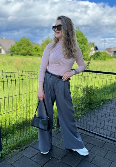 Top de manga larga rosa claro, pantalones de pierna ancha grises, bolso negro y zapatillas blancas. El fondo presenta hierba verde y un cielo nublado.