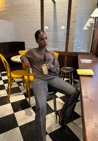 Frau in brauner Bluse und grauen Jeans sitzt auf einem Hocker in einem Café und hält ein Getränk mit einem Strohhalm. Gelbe Stühle und ein schwarz-weißer Fliesenboden sind sichtbar.