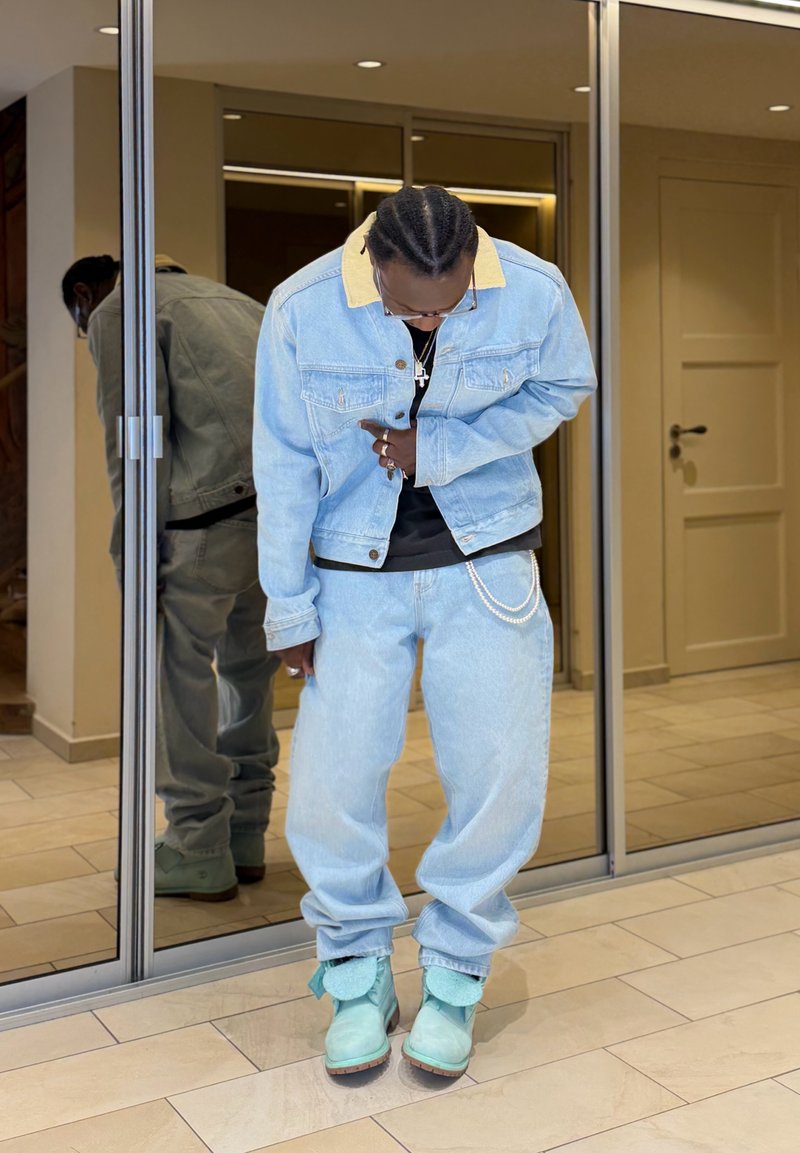 Homme portant une veste en jean bleu clair et un pantalon avec des bottes turquoise, debout à l'intérieur devant des portes coulissantes en verre, la tête baissée.