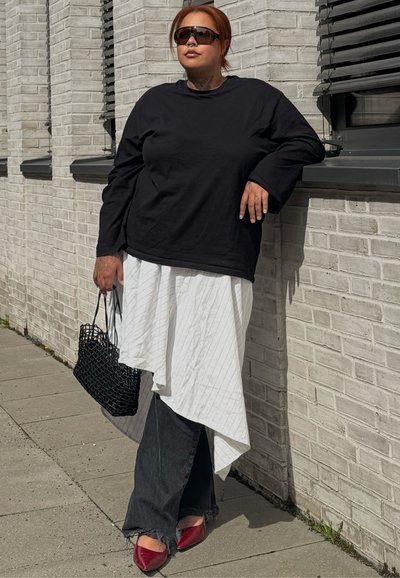 Mujer con cabello rojo y gafas de sol se apoya en una pared clara de ladrillo sosteniendo un bolso negro tejido, vistiendo una blusa negra, falda blanca, pantalones negros y zapatos rojos.