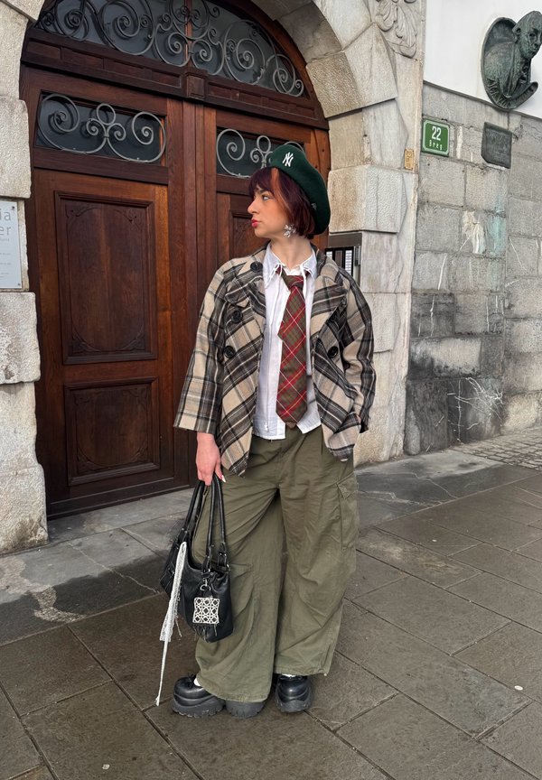 Mladý člověk s zeleným baretíkem, kostkovaným sakem, červenou kostkovanou kravatou, bílou košilí, širokými olivovými kalhotami a botami na platformě stojí na kamenné dlažbě u dřevěných dveří.