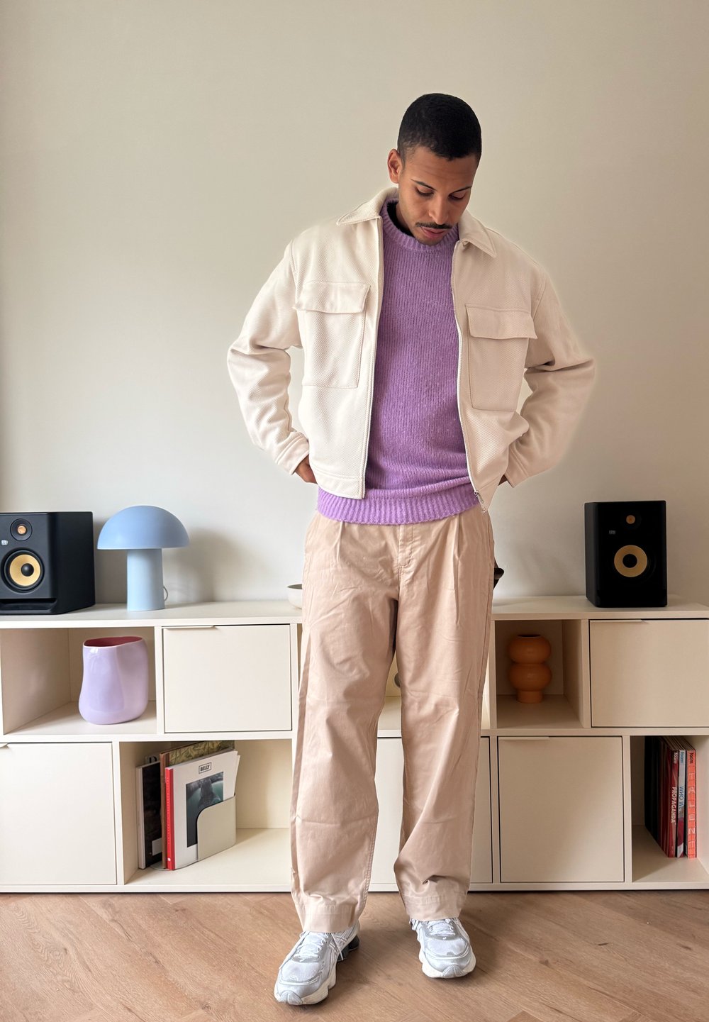 Kremfarget jakke med to frontlommer, lys lilla genser, beige bukser og grå joggesko. Bakgrunnen har minimalistiske møbler og dekor.