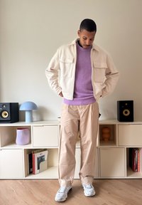 Kremfarget jakke med to frontlommer, lys lilla genser, beige bukser og grå joggesko. Bakgrunnen har minimalistiske møbler og dekor.