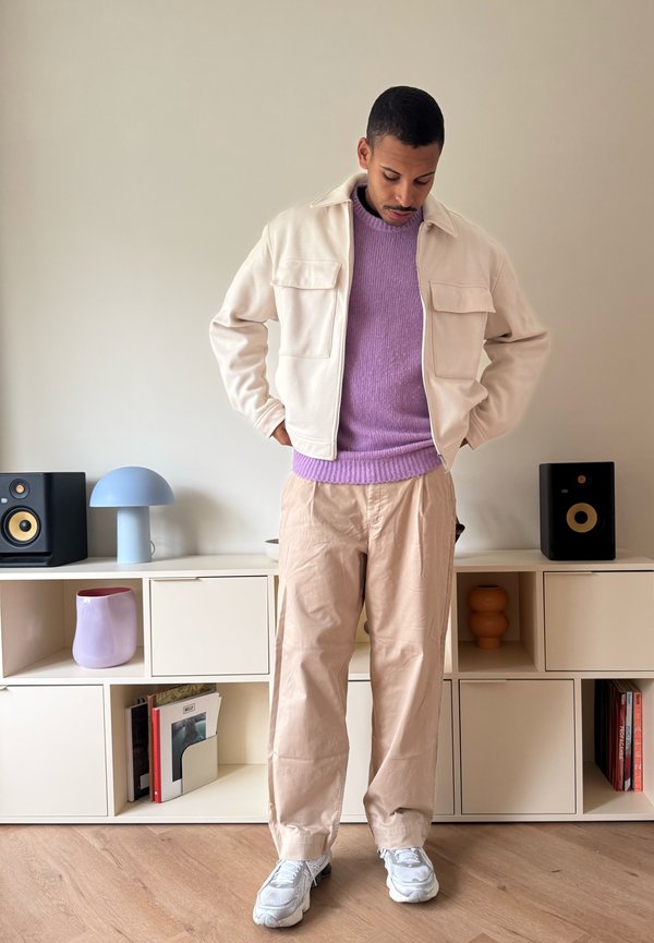 Crèmekleurig jack met twee voorzakken, lichtpaarse trui, beige broek en grijze sneakers. De achtergrond toont minimalistische meubels en decoratie.