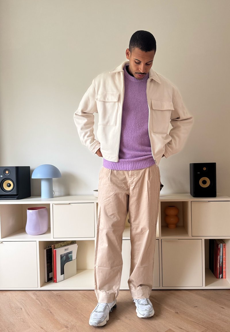 Casaco creme com dois bolsos frontais, suéter roxo claro, calças bege e ténis cinzentos. O fundo apresenta móveis e decoração minimalistas.