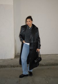 Sort læder trenchcoat over en grå sweater, kombineret med lyseblå jeans og sorte sko. Tilbehør inkluderer en lille sort håndtaske.