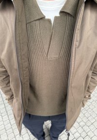 Mężczyzna w beżowej kurtce na zamek błyskawiczny, pod którą ma beżowy, prążkowany sweter, oraz w granatowych spodniach, stoi na jasnoszarej, kafelkowanej nawierzchni.