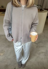 Personne en pull beige côtelé à fermeture éclair et pantalon blanc tenant un gobelet en papier avec un café mousseux à l'intérieur sur un sol en béton.