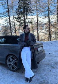 Homme portant des lunettes de soleil, un long manteau, une écharpe et des gants, appuyé sur une voiture décapotable grise dans une forêt enneigée avec de grands arbres et des montagnes en arrière-plan.