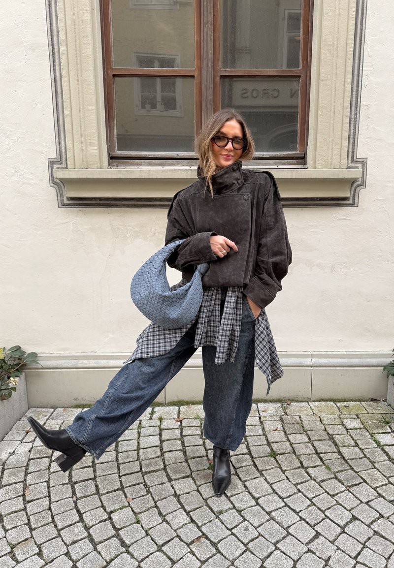 Femme en veste sombre surdimensionnée, chemise à carreaux nouée à la taille, jeans larges bleu, bottes noires et lunettes tenant un sac tissé bleu dans une rue pavée.