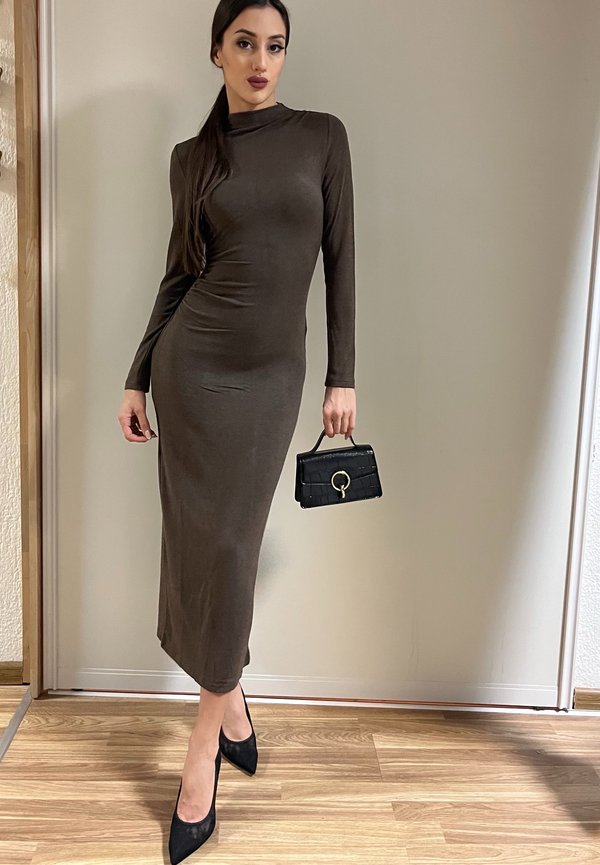 Nainen poseeraa istuvassa, pitkähihnaisessa ruskeassa midi-mekossa, kädessään pieni musta käsilaukku ja jalassaan mustat teräväkärkiset korkokengät puisella lattialla.