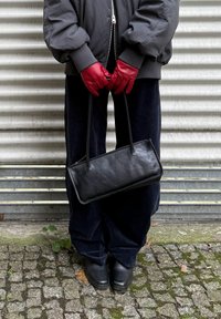 Borsa nera in pelle con manici lunghi, forma rettangolare, tenuta da mani guantate, indossata con pantaloni scuri e larghi e una giacca.