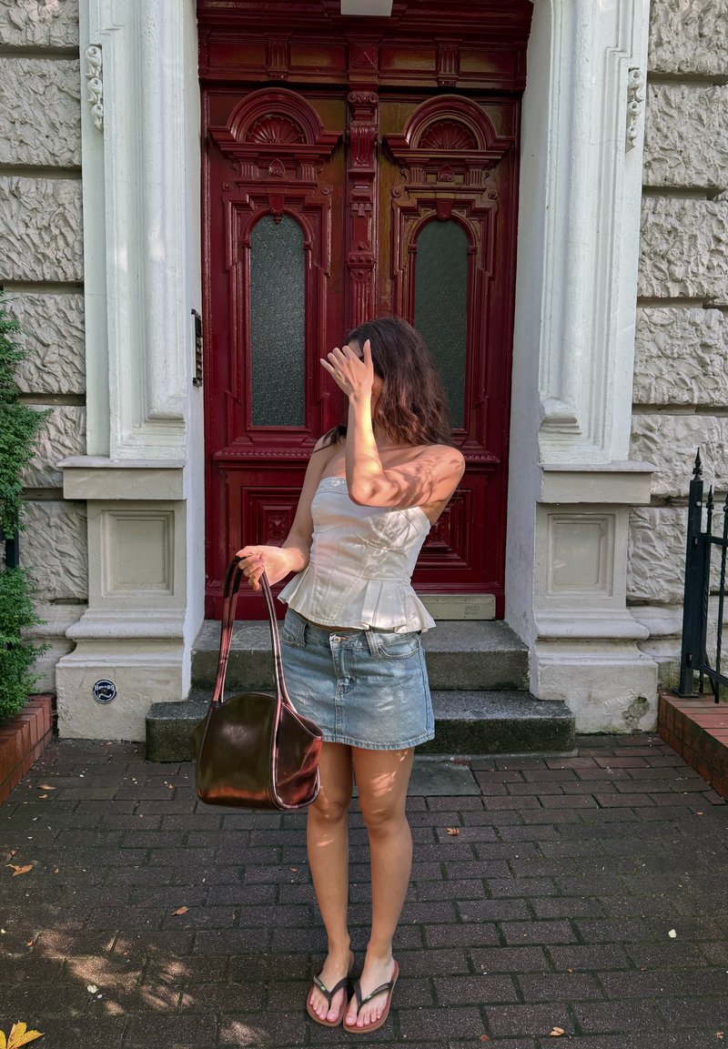 Femme en haut blanc bustier et jupe en jean tenant un sac marron, se protégeant le visage avec la main, debout devant une grande porte rouge.