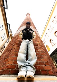 Jeune homme portant une veste noire et crème et un jean ample, debout les mains dans les poches contre une haute cheminée en briques rouges entre des bâtiments.