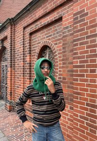 Osoba koja nosi sunčane naočale i zeleni šal s kapuljačom, odjevena u prugasti pulover i traperice, stoji ispred zidane crvene cigle s ukrasnim prozorima.